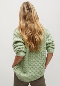 Vaaleanvihreä neulottu neule, jossa on kuvioituja kuvioita, pudonneet hartiat ja resorit hihansuissa. Päällä kuvioidut housut, kuvattu takaa päin.