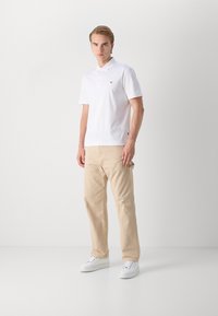 Camisa pólo branca de mangas curtas e três botões, combinada com calças bejas de perna reta e ténis brancos, num fundo neutro.