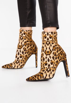 Bottes à talons aiguilles à imprimé léopard montant à la cheville, avec des bouts pointus et des fermetures éclair latérales, portées avec un pantalon noir raccourci.