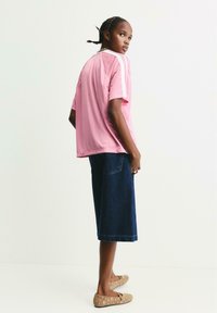 Unge person med fletninger iført en løs lyserød bluse med hvide striber, mørk denim shorts med bred benvidde og beige flade sko med hulmønster.