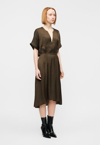 Φόρεμα ελιάς πράσινο με βαθύ V λαιμόκοψη, κοντά μανίκια και ελαστική μέση. Ρούχο με ρέον ύφασμα, μήκος ως τη μέση της γάμπας, συνδυασμένο με μαύρα μποτάκια για τον αστράγαλο.