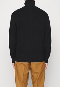 Maglione nero a coste con colletto alto, caratterizzato da maniche lunghe e una vestibilità ampia, abbinato a pantaloni beige a gamba larga.