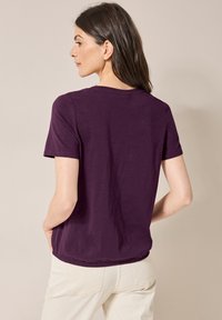 T-shirt a maniche corte di un profondo viola, tessuto morbido con colletto a girocollo. Presenta una vestibilità rilassata e un orlo curvo, mostrato dal retro.