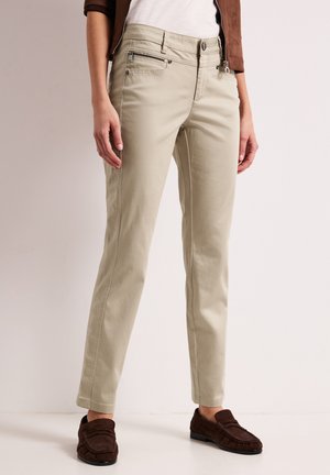Kvinde iført beige slim-fit bukser med for- og møntlommer, kombineret med brune loafers og en brun jakke over en hvid top.