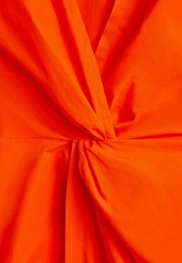 Tissu orange à texture lisse, présentant un drapé torsadé au centre. La couleur est vive et audacieuse, renforçant l'impact visuel du design.