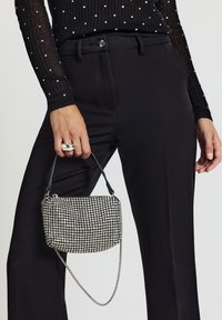 Kleine Handtasche mit einer strukturierten silbernen Oberfläche, die ein Gitter aus glänzenden Akzenten aufweist. Schwarzer Griff und Kettenriemen. Kombiniert mit schwarzen Hosen mit weitem Bein.