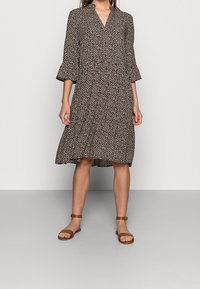 Tryckt klänning med v-ringning, trekvarts ärmar med klockform, åtdragen midja och knälång kjol. Färgen har en svart bas med kontrasterande små mönster. Bruna sandaler.