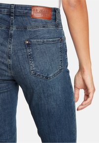 Denimjeans med bakficka och en brun läderlogga, som bärs av en person med synlig högra hand och handled.
