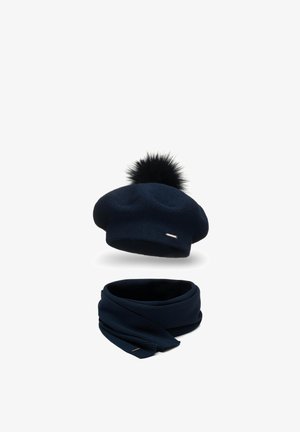 Marine gebreide baret met een zwarte nepbont pompon en een bijpassende infinity sjaal. Beide stukken hebben een klein, glanzend metaalelement.