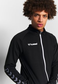 Sort zip-jakke med et tekstureret stof, der har en hvid frontlynlås og hummel-logo, samt kontrasterende stribede detaljer langs ærmerne.