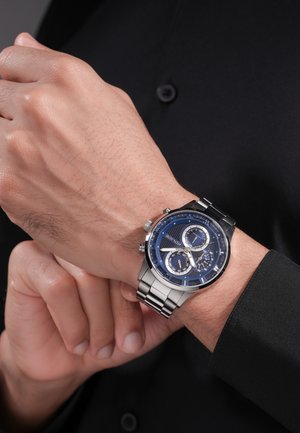 Orologio da polso con quadrante blu e cinturino in metallo argento indossato al polso sinistro, mostrato mentre si regola o controlla l'orologio con la mano opposta.