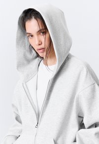 Grå hettejakke med glidelås og stor hette, laget av mykt bomullsmateriale, med frontlomme og ribbestrikkede mansjetter. Glatt tekstur, avslappet passform.