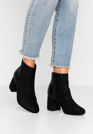 Bottes en suède noir longueur cheville avec talons blocs et fermetures éclair, portées avec un jean bleu clair à ourlet effiloché.