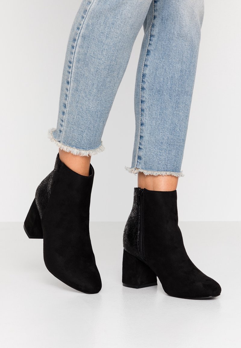 Bottes en suède noir longueur cheville avec talons blocs et fermetures éclair, portées avec un jean bleu clair à ourlet effiloché.