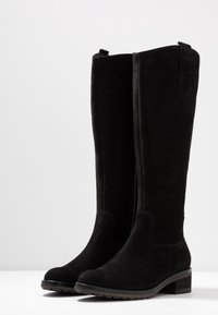 Un par de botas de ante negras hasta la rodilla con tacones bajos y punteras redondeadas, de pie sobre una superficie blanca.
