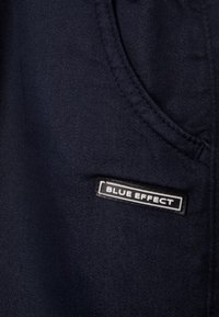 Tmavě modrá látka s hladkým povrchem má boční kapsu a obdélníkovou etiketu "BLUE EFFECT" v bílé a černé barvě.