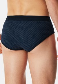 Navy modré slipové spodní prádlo vyrobené z texturované látky s geometrickým vzorem, s černým elastickým pasem a nízkými průramky.