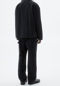 Black outerwear jacket with a relaxed fit, paired with loose-fitting black cargo pants, featured from the back. Smooth cotton texture.