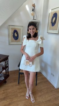 Žena u bijeloj mini haljini s rukavima u obliku mašne, zlatnim naušnicama u obliku cvijeta i odgovarajućom torbicom stoji i smiješi se u sobi s drvenim podom i uokvirenom plavom umjetninom.