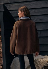 Brun fleecejakke med grå krave, afslappet pasform og lynlåsdetalje i siden. Bruges over sorte bukser mod en mørk træbaggrund.