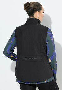 Schwarze ärmellose Weste mit hohem Kragen, ausgestattet mit einer glatten Textur und einem dezenten Branding-Detail. Getragen über einem gemusterten Langarmhemd.