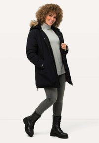 Svart isolerad parka med en fuskpälsfodrad huva, dragkedja framtill och sidofickor, kombinerad med en grå stickad tröja och grå jeans. Svarta kängor.