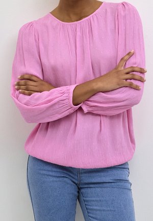 Person wearing a textured long-sleeve pink blouse with arms crossed, paired with light blue fitted jeans against a plain background.