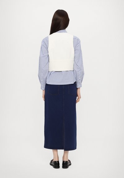 Hvit tettsittende vest over en blå gingham-skjorte, kombinert med et langt marineblått denimskjørt med splitt midt bak og svarte sko.