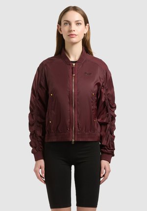 Woman wearing a maroon bomber jacket with ruched sleeves and gold zipper, paired with black knee-length fitted shorts, standing against a plain background.