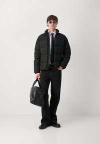 Jachetă puffer neagră, asociată cu o cămașă albă și o cravată neagră. Modelul ține o geantă neagră, poartă ochelari de soare și pantaloni negri. Textură netedă.