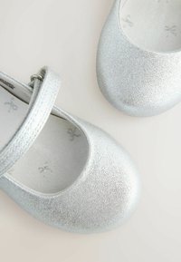 Sudraba mirdzoši Mary Jane stila apavi ar apaļu purngalu, ar siksniņu un sprādzi, kā arī teksturētu augšdaļu. Oderēti ar loku rakstu.