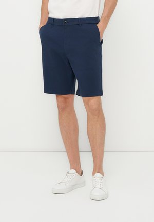 Mężczyzna ubrany w granatowe, dopasowane krótkie spodenki z bocznymi kieszeniami, białe sneakersy i białą koszulę, stojący na tle jednolitego białego tła.