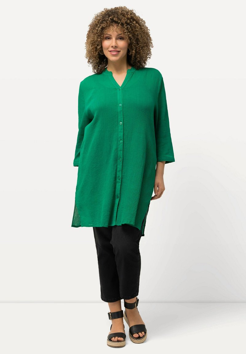 Camisa verde de botones con escote en V, mangas tres cuartos y aberturas laterales, combinada con pantalones negros cortos y sandalias de plataforma negras.