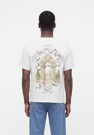 Homme portant un t-shirt blanc avec un arc architectural et texte en espagnol au dos, associé à un jean bleu, debout de dos.