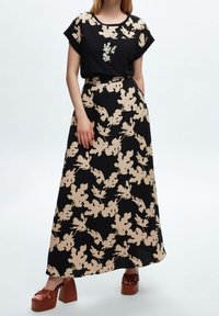 Vestido negro con patrón de hojas beige, mangas cortas y cintura definida, combinado con sandalias de plataforma marrones con un diseño de tiras cruzadas.