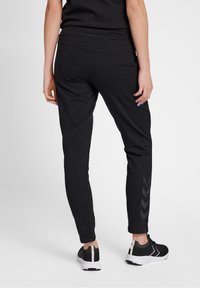 Svarta träningsoveraller med en smal passform, med en bred midjeband och en subtil chevronmönster längs nederdelen av benet. Bärs med svarta sneakers.