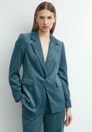Frau mit langen, glatten Haaren, die eine tealfarbene Cordjacke und passende Hose trägt, steht vor einem schlichten weißen Hintergrund.