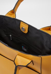 Żółta torebka tote z teksturowanej skóry z czarnym wnętrzem, wyposażona w kieszeń na zamek i zapięcie na zatrzask. Detale obejmują kontrastowe przeszycia na krawędziach.