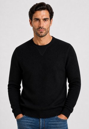 Mand med mørkt hår og skæg iført en sort tekstureret crewneck sweater og blå jeans, stående med hænderne i lommerne mod en ensfarvet baggrund.