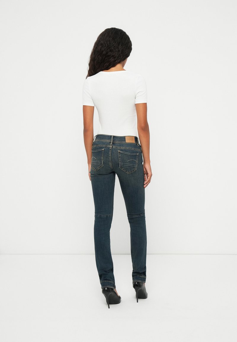 Donkerblauwe jeans met een slanke pasvorm, gekenmerkt door een yoke en zichtbare stiksels, gecombineerd met een witte aansluitende top en hoge hakken.