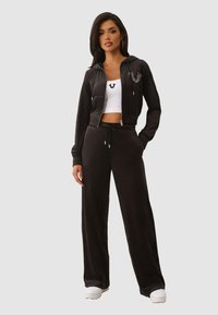 Schwarzer Velours-Trainingsanzug mit einem kurzen, zip-up Hoodie mit Logo, kombiniert mit locker sitzenden Hosen. Das Model trägt ein weißes Crop-Top und Sneakers.