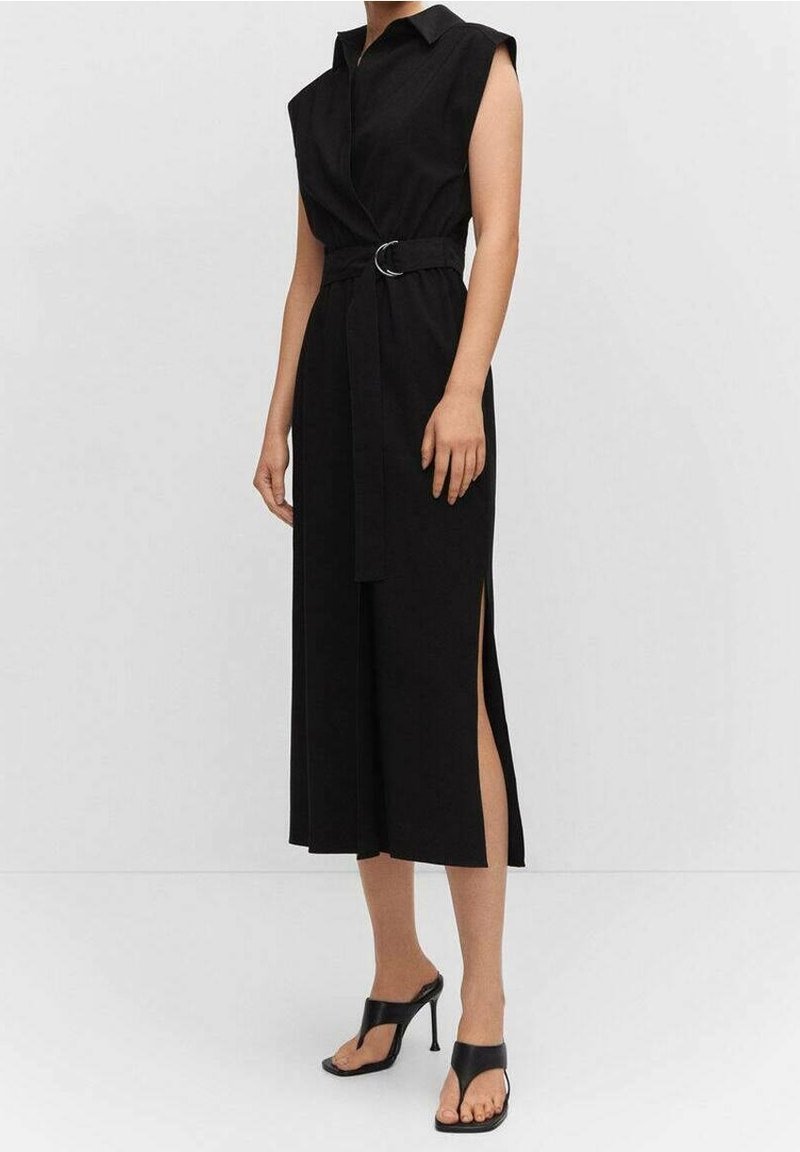 Femme portant une robe midi noire sans manches ceinturée avec un col et une fente latérale, associée à des sandales noires à talons hauts.