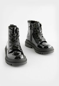 Bottes noires brillantes à lacets avec semelles épaisses et fermetures éclair latérales, dotées de lacets en tissu doux et d'intérieurs doublés de fourrure, présentées sur un fond blanc.