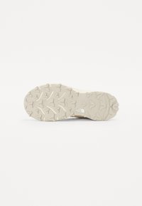 Semelle de chaussure beige clair avec un motif de grip texturé, comportant des crampons triangulaires pour l'adhérence et un logo de The North Face.