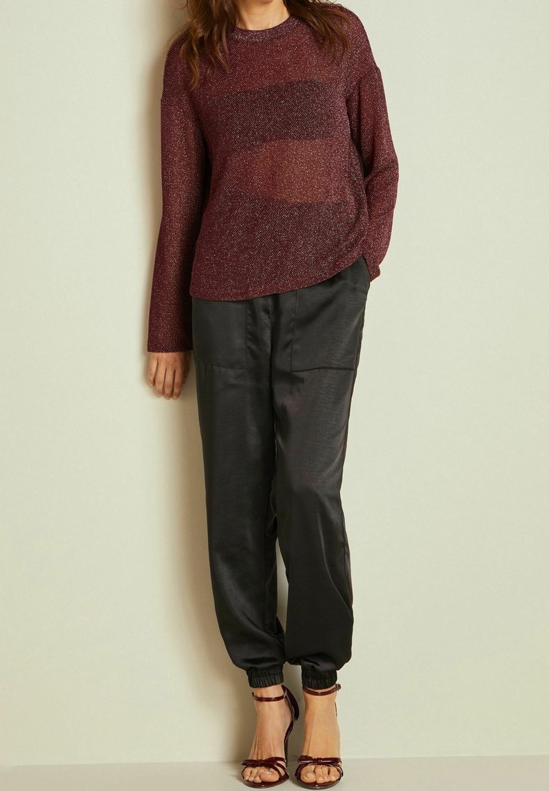 Femme portant un haut transparent manches longues bordeaux, un pantalon de jogging en satin noir avec des poches, et des sandales à talons hauts bordeaux, debout contre un mur uni.