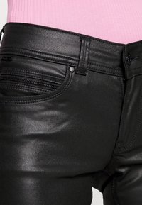 Mustat, venyvät tekonahkahousut, joissa on sileä pinta, vetoketjullinen tasku ja vartalonmyötäinen muotoilu, yhdistettynä vaaleanpunaiseen ribbipaitaan.