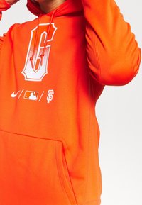 Sweat à capuche orange avec un grand logo graphique "G" blanc, arborant les marques MLB et San Francisco, et une poche avant pour le rangement.