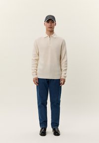 Mann trägt einen cremefarbenen, gerippten Pullover mit Reißverschluss am Kragen, blaue Jeans, schwarze Schuhe und eine dunkle Baseballkappe, steht vor einem schlichten hellen Hintergrund.