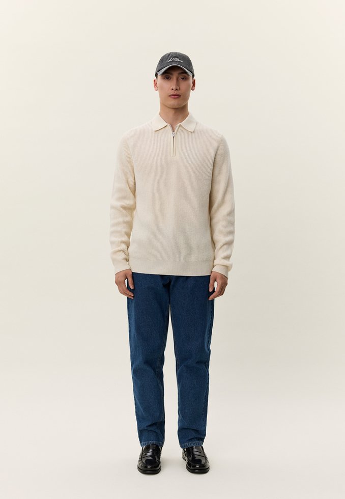 Mann trägt einen cremefarbenen, gerippten Pullover mit Reißverschluss am Kragen, blaue Jeans, schwarze Schuhe und eine dunkle Baseballkappe, steht vor einem schlichten hellen Hintergrund.