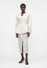 Vrouw draagt een gestructureerde witte blazer en bijpassende midi-rok met splits aan de voorkant, gecombineerd met zwarte sandalen met hak, staand tegen een effen achtergrond.